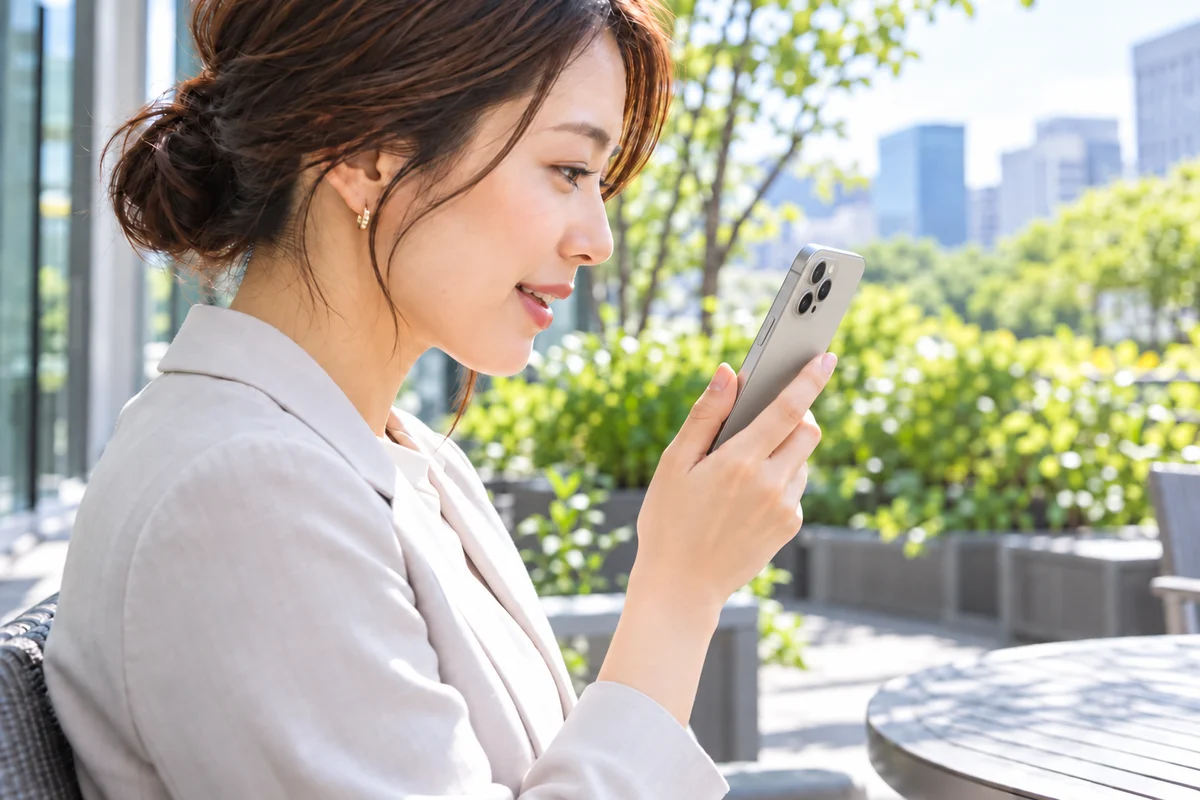 屋外のテラスで、日本人女性がスマホに話しかけてホームページの更新を指示しているイメージ
